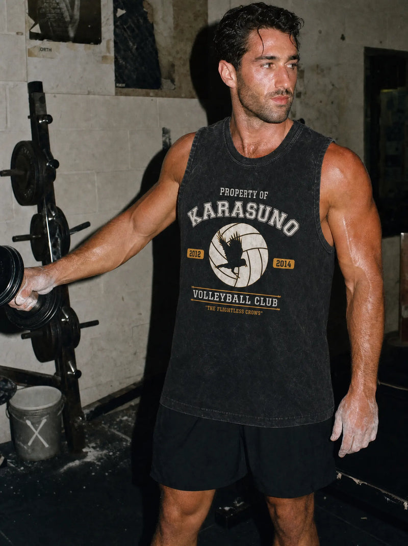 A muscular man in a gym wearing a dark charcoal gray graphic tank top that reads 'PROPERTY OF KARASUNO VOLLEYBALL CLUB' with a volleyball graphic, paired with black athletic shorts.